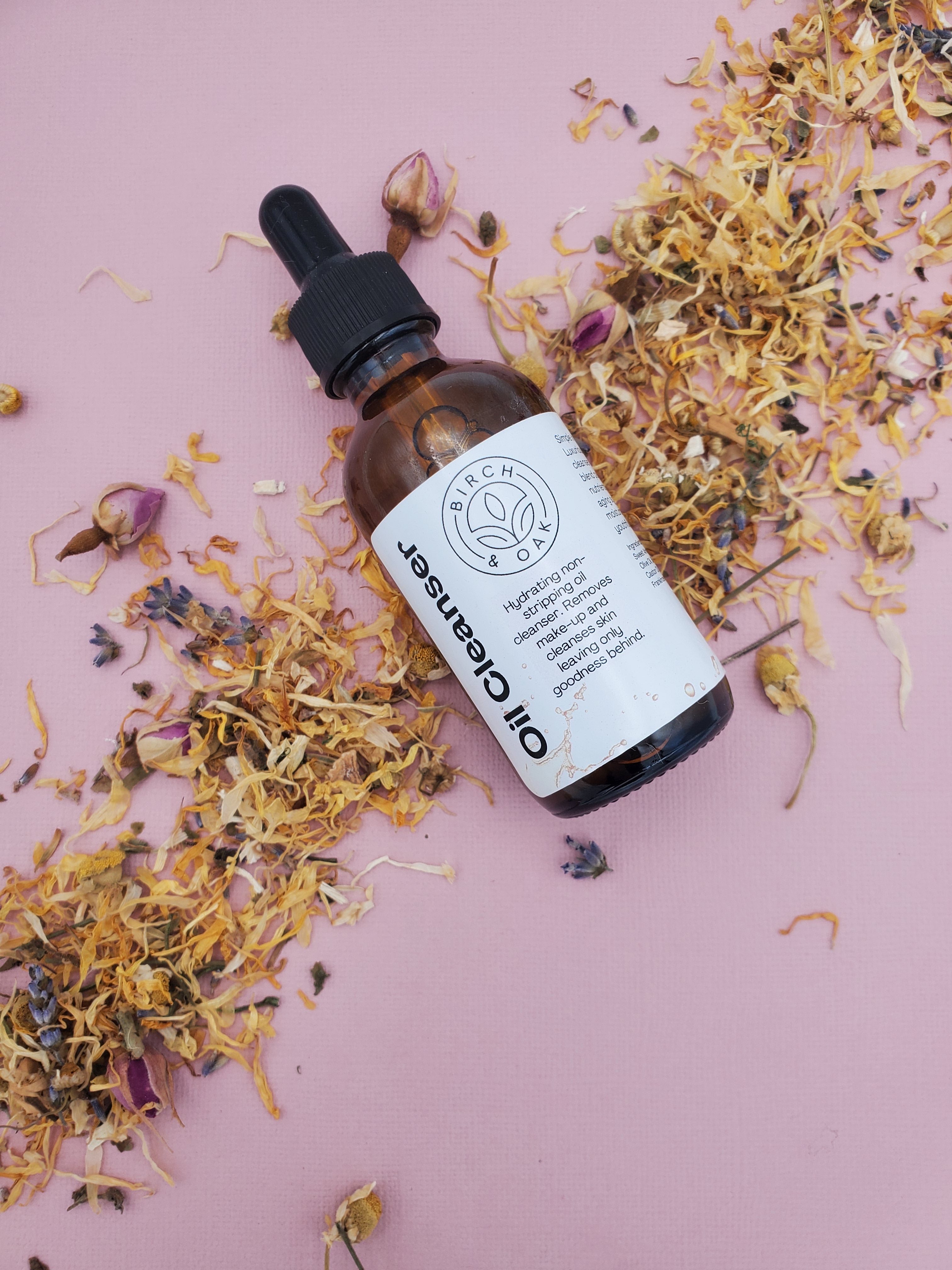 A bottle of Facial Oil Cleanser with a dropper top, surrounded by dried floral petals, presented on a pink background.