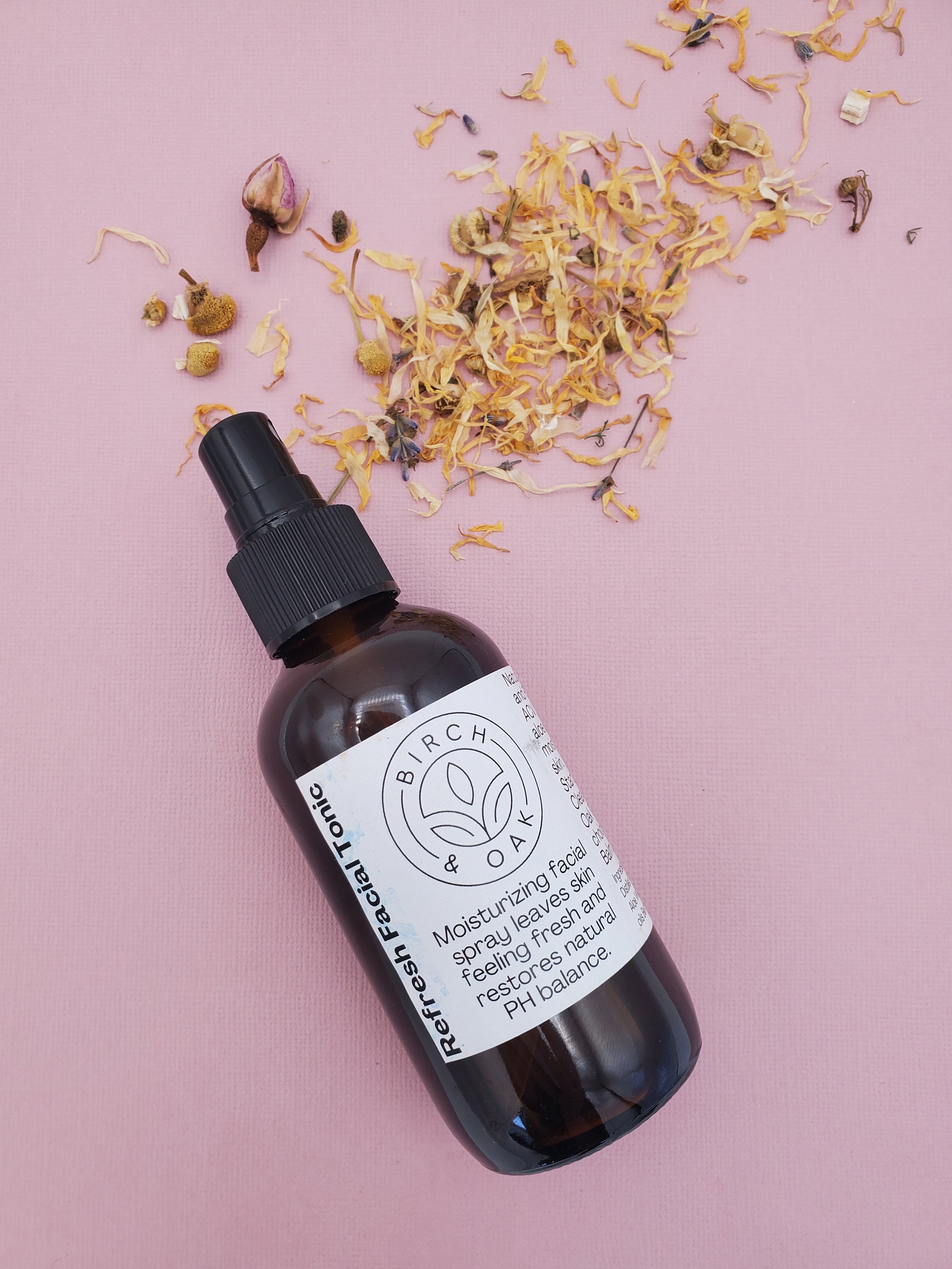 A bottle of Refresh PH Balancing Toner with a dropper cap, surrounded by dried floral petals, presented on a pink background.
